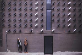 Two individuals standing in front of a wall covered with various security cameras, showcasing an urban art installation.