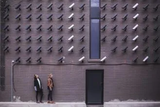 Two individuals standing in front of a wall covered with various security cameras, showcasing an urban art installation.