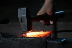 A blacksmith's hand holding a hammer, striking a piece of glowing red-hot metal on an anvil, with sparks flying.