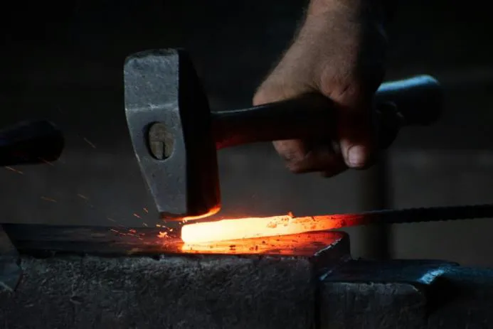A blacksmith's hand holding a hammer, striking a piece of glowing red-hot metal on an anvil, with sparks flying.