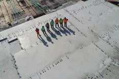 Aerial view of a construction site with a team of six workers in safety gear standing on a concrete slab, surrounded by construction materials and tools.