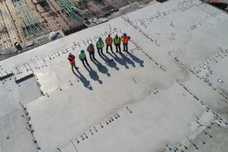 Aerial view of a construction site with a team of six workers in safety gear standing on a concrete slab, surrounded by construction materials and tools.