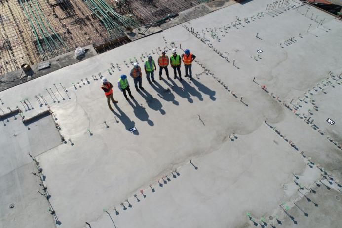 Aerial view of a construction site with a team of six workers in safety gear standing on a concrete slab, surrounded by construction materials and tools.