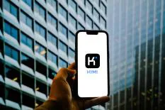 A hand holding a smartphone displaying the Kimi logo in front of modern glass buildings.