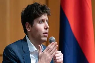 A man in a blue suit speaking into a microphone at a conference, with a flag in the background.